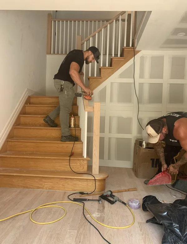 Craftsman sanding new oak staircase treads beside picture-frame wainscoting